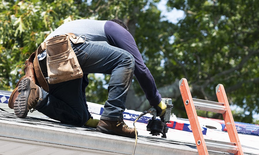 A Contractor Is Kneeling On A Residential Roof Using A Nailgun T Roof Replacements Near Me